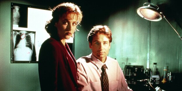 Dana Scully und Fox Mulder aus der Serie "Akte-X" blicken in die Kamera. Im Hintergrund hängen Röntgenbilder Dana Scully und Fox Mulder aus der Serie "Akte-X" blicken in die Kamera. Im Hintergrund hängen Röntgenbilder