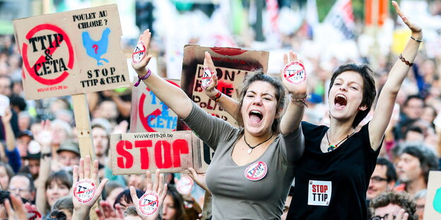 Zwei frauen reißen die Arme in die Luft. Dahinter Schilder mit "Stopp TTIP"