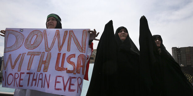 Eine Frau hält ein Plakat, auf dem steht „Down with USA. More than ever“, zwei andere heben die Hand in die Höhe Eine Frau hält ein Plakat, auf dem steht „Down with USA. More than ever“, zwei andere heben die Hand in die Höhe