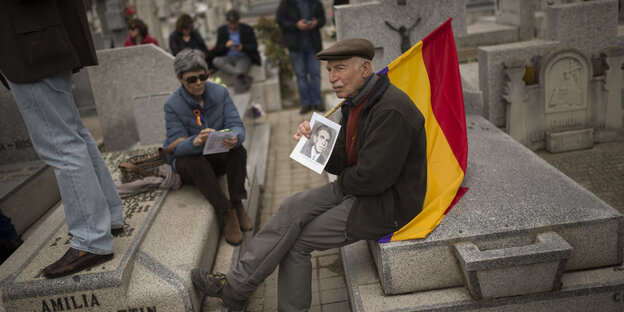 Ein Mann sitzt auf einem Grabstein und trägt die Fahne der spanischen 2. Republik über der Schulter, in der Hand hält er eine schwarz-weiß Fotografie Ein Mann sitzt auf einem Grabstein und trägt die Fahne der spanischen 2. Republik über der Schulter, in der Hand hält er eine schwarz-weiß Fotografie