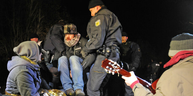 Zwei Polizisten tragen einen Aktivisten weg, zwei Aktivisten sitzen daneben, einer davon spielt Guitarre Zwei Polizisten tragen einen Aktivisten weg, zwei Aktivisten sitzen daneben, einer davon spielt Guitarre