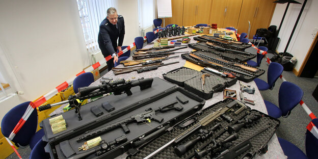 Auf einem langen Tisch liegen verschiedene Arten von Waffen. Im Hintergrund ein Mann in Polizeikleidung. Auf einem langen Tisch liegen verschiedene Arten von Waffen. Im Hintergrund ein Mann in Polizeikleidung.
