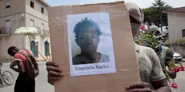 Maskierter Mann hält Karton mit Foto von Sacko Soumali in die Kamera Maskierter Mann hält Karton mit Foto von Sacko Soumali in die Kamera