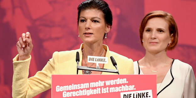 ein Frau mit erhobener Hand an einem Rednerpult, rechtas daneben eine andere Frau ein Frau mit erhobener Hand an einem Rednerpult, rechtas daneben eine andere Frau
