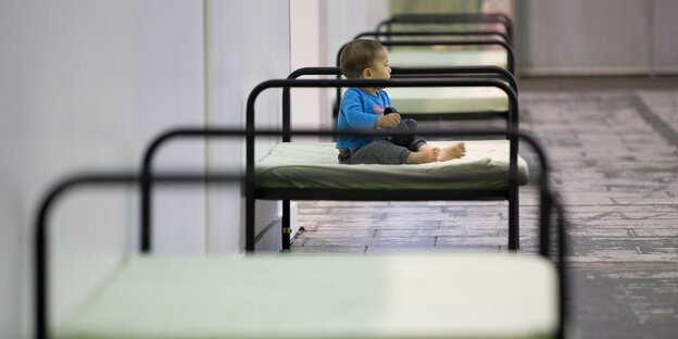 In Hangar 2 des ehemaligen Flughafen Tempelhof: Kleines Kind sitzt auf einem spartanischen Bett In Hangar 2 des ehemaligen Flughafen Tempelhof: Kleines Kind sitzt auf einem spartanischen Bett