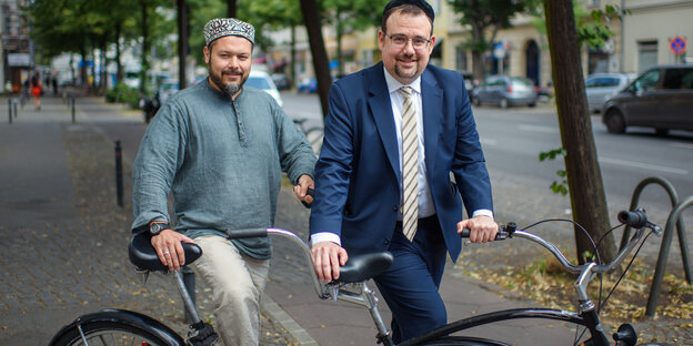 Vor einem großen Tandem-Fahrrad steht ein Mann mit einer muslimischen Kopfbedeckung, daneben ein Mann mit einer jüdischen Kopfbedeckung Vor einem großen Tandem-Fahrrad steht ein Mann mit einer muslimischen Kopfbedeckung, daneben ein Mann mit einer jüdischen Kopfbedeckung