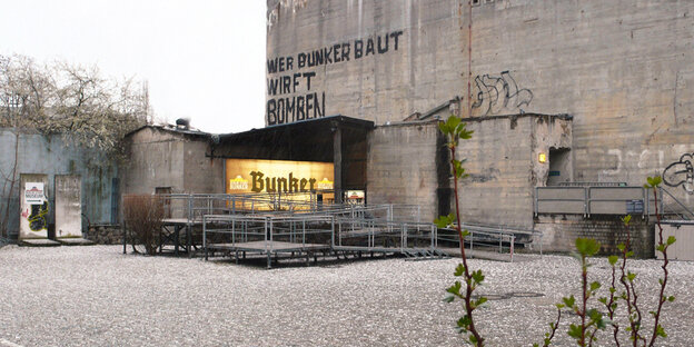 Ein Hof und ein Bunker. Auf dem Bunker steht ein Graffito: "Wer Bunker baut, wirft Bomben." Ein Hof und ein Bunker. Auf dem Bunker steht ein Graffito: "Wer Bunker baut, wirft Bomben."
