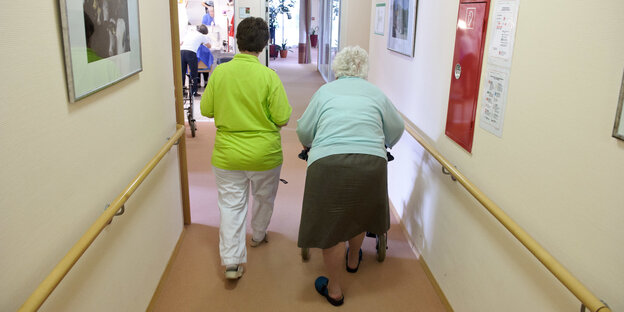 Eine Pflegerin und eine alte Frau mit Rollator gehen über einen Flur Eine Pflegerin und eine alte Frau mit Rollator gehen über einen Flur