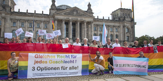 Eine Menschenmenge steht vor dem Bundestag. Sie halten eine Regenbogenflagge und Banner hoch. Die Menschen lächeln in die Kamera. Eine Menschenmenge steht vor dem Bundestag. Sie halten eine Regenbogenflagge und Banner hoch. Die Menschen lächeln in die Kamera.