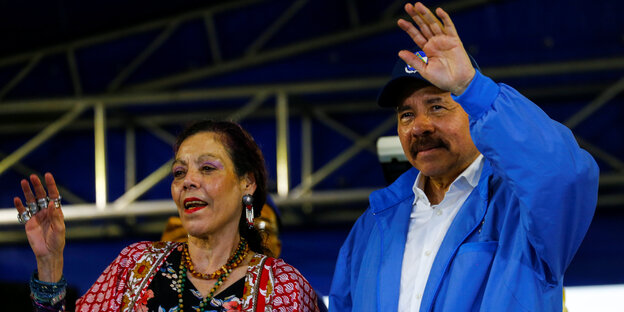 Rosario Murillo, in buntem Kleid und mit vielen Ringen an der Hand, und Daniel Ortega in blauer Strickjacke winken von einer Bühne. Rosario Murillo, in buntem Kleid und mit vielen Ringen an der Hand, und Daniel Ortega in blauer Strickjacke winken von einer Bühne.
