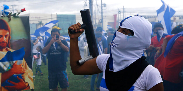 Im Vordergrund eine maskierte Demonstrantin, die einen selbstgebauten Granatwerfer in der Hand hält. Im Hintergrund Menschen mit blau-weißen nicaraguanischen Fahnen. Im Vordergrund eine maskierte Demonstrantin, die einen selbstgebauten Granatwerfer in der Hand hält. Im Hintergrund Menschen mit blau-weißen nicaraguanischen Fahnen.