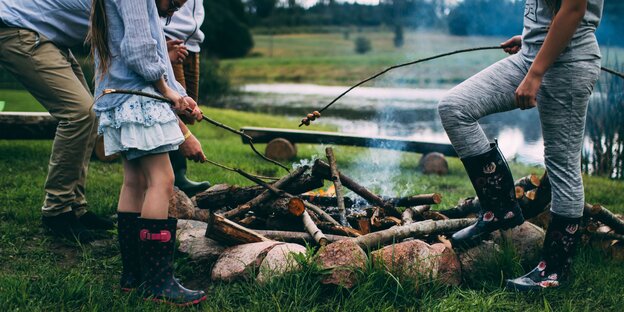 Mehrere Personen stehen an einem Lagerfeuer und grillen Lebensmittel auf Stöcken Mehrere Personen stehen an einem Lagerfeuer und grillen Lebensmittel auf Stöcken