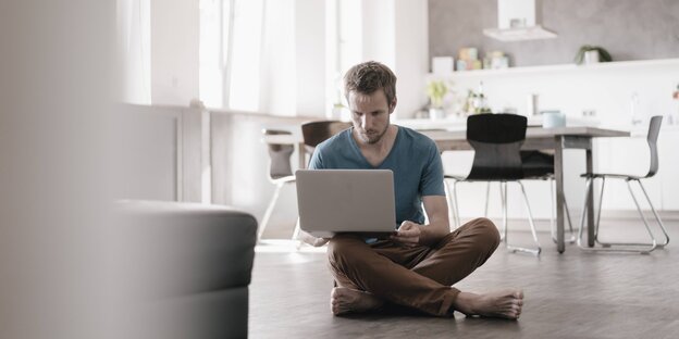 Ein Mann sitzt mit einem Laptop auf dem Boden in einer offenen Küche Ein Mann sitzt mit einem Laptop auf dem Boden in einer offenen Küche