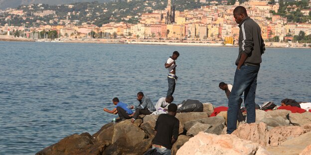 Menschen sitzen und stehen auf Steinen am Meer, im Hintergrund sieht man die italienische Stadt Ventimiglia Menschen sitzen und stehen auf Steinen am Meer, im Hintergrund sieht man die italienische Stadt Ventimiglia
