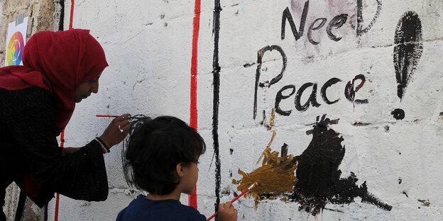 Eine Frau und ein kleiner Junge malen etwas an eine Wand. Zu lesen ist schon: "Need Peace" Eine Frau und ein kleiner Junge malen etwas an eine Wand. Zu lesen ist schon: "Need Peace"
