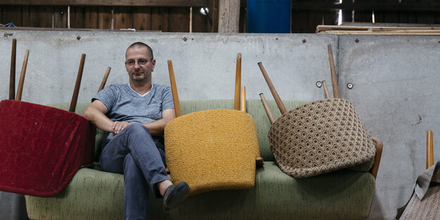 Ein Mann sitzt mit verschränkten Armen auf einem Sofa, neben ihm liegen Stühle auf dem Sofa Ein Mann sitzt mit verschränkten Armen auf einem Sofa, neben ihm liegen Stühle auf dem Sofa