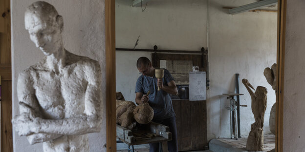 Ein Mann arbeitet in einem Atelier an einem Kunstwerk aus Holz, im Vordergrund steht eine Statue Ein Mann arbeitet in einem Atelier an einem Kunstwerk aus Holz, im Vordergrund steht eine Statue
