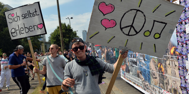 Junger Mann mit Sonnenbrille grinst und hält Schild hoch, darauf sind zwei Herzen, ein Peace-Zeichen und Noten zu sehen Junger Mann mit Sonnenbrille grinst und hält Schild hoch, darauf sind zwei Herzen, ein Peace-Zeichen und Noten zu sehen