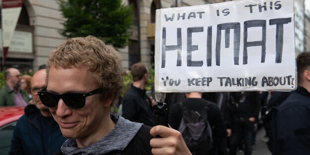 Ein Mann mit Sonnenbrille hält ein Plakat mit der Aufschrift „What is this Heimat you keep talking about“ hoch Ein Mann mit Sonnenbrille hält ein Plakat mit der Aufschrift „What is this Heimat you keep talking about“ hoch