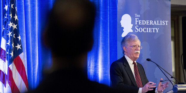 John Bolton steht an einem Rednerpult, vorne im Bild ist als dunkle Silhouette ein Mann von hinten zu sehen John Bolton steht an einem Rednerpult, vorne im Bild ist als dunkle Silhouette ein Mann von hinten zu sehen