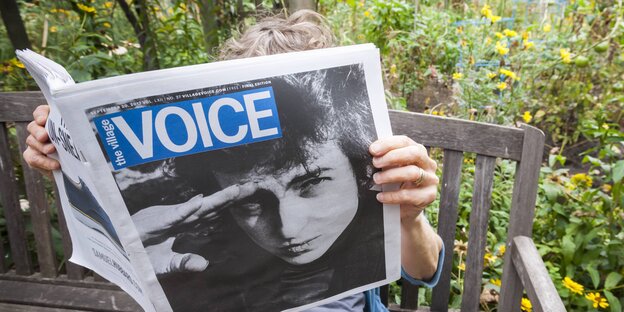 Eine Person sitzt auf einer Bank und hält eine Ausgabe der Zeitung "Village Voice" in der Hand Eine Person sitzt auf einer Bank und hält eine Ausgabe der Zeitung "Village Voice" in der Hand