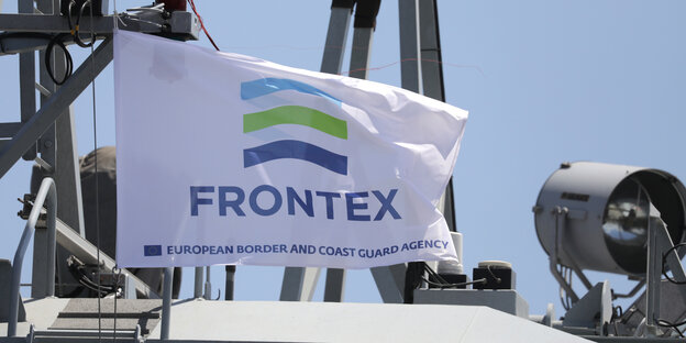 Eine weiße Flagge, auf der Frontex steht, im Hintergrund ein Schiff und blauer Himmel Eine weiße Flagge, auf der Frontex steht, im Hintergrund ein Schiff und blauer Himmel