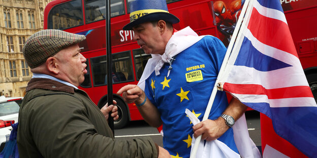Zwei Männer diskutieren und gestikulieren. Einer von ihnen trägt eine EU- und eine britische Flagge Zwei Männer diskutieren und gestikulieren. Einer von ihnen trägt eine EU- und eine britische Flagge