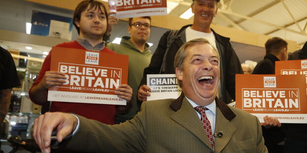 Nigel Farage lacht, hinter ihm stehen Männer mit Plakaten Nigel Farage lacht, hinter ihm stehen Männer mit Plakaten