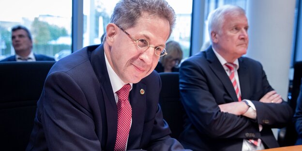 ein Mann schmunzelt am Tisch sitzend. Im Hintergrund mit verschränkten Armen: Horst Seehofer ein Mann schmunzelt am Tisch sitzend. Im Hintergrund mit verschränkten Armen: Horst Seehofer