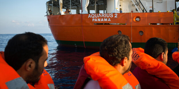 Drei Männer in orangefarbenen Schwimmwesten blicken von einem Boot auf ein Schiff Drei Männer in orangefarbenen Schwimmwesten blicken von einem Boot auf ein Schiff