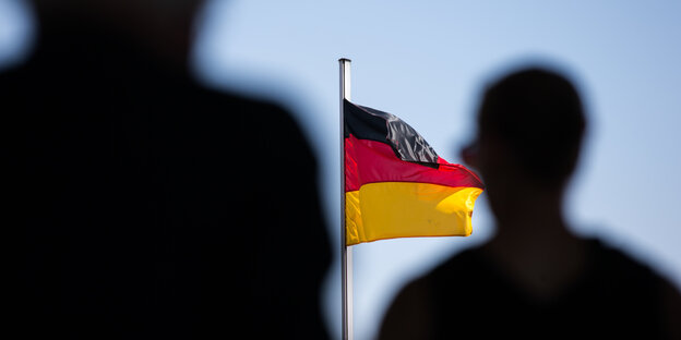 Eine Deutschlandfahne weht an einem Fahnenmast, davor stehen zwei Menschen. Eine Deutschlandfahne weht an einem Fahnenmast, davor stehen zwei Menschen.