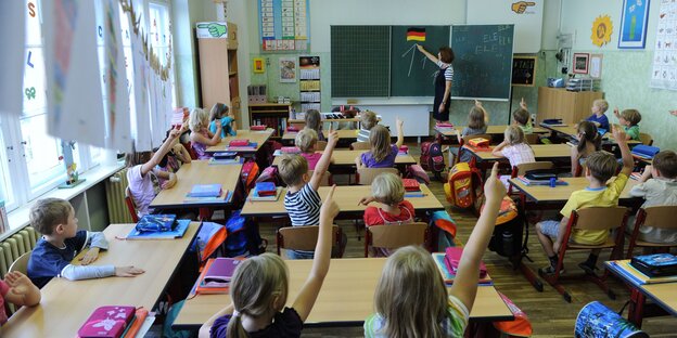 Erstklässler sitzen in einem Klassenraum und melden sich. Vorne steht die Lehrerin an der Tafel.