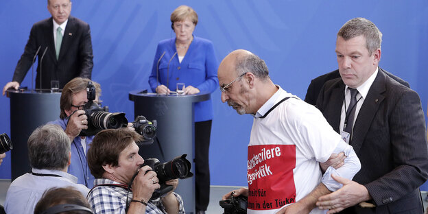 Vor einem Mann sind viele Fotografen. Der Mann wird von einem anderen hinter ihm an den Armen festgehalten Vor einem Mann sind viele Fotografen. Der Mann wird von einem anderen hinter ihm an den Armen festgehalten