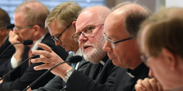 Mehrere Männer sitzen hinter Mikrofonen, einer spricht Mehrere Männer sitzen hinter Mikrofonen, einer spricht