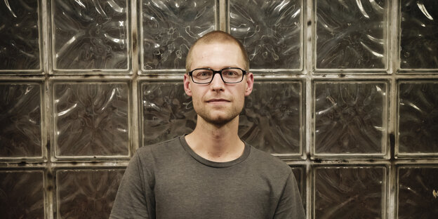 Das Bild zeigt einen schlanken, jungenhaft wirkenden Mann mit Brille und sehr kurz geschnittenen blonden Haaren in einem grauen T-Shirt vor einer Wans aus Glasbausteinen. Das Bild zeigt einen schlanken, jungenhaft wirkenden Mann mit Brille und sehr kurz geschnittenen blonden Haaren in einem grauen T-Shirt vor einer Wans aus Glasbausteinen.