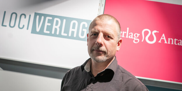 Ein Mann mit kurzen Haaren vor einer Wand mit der Aufschrift "Loci-Verlag" Ein Mann mit kurzen Haaren vor einer Wand mit der Aufschrift "Loci-Verlag"