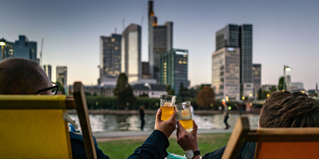 Zwei Menschen in Liegestühlen sind von hinten zu sehen, sie prosten sich mit Appelwoi zu und blicken auf die Skyline von Frankfurt am Main