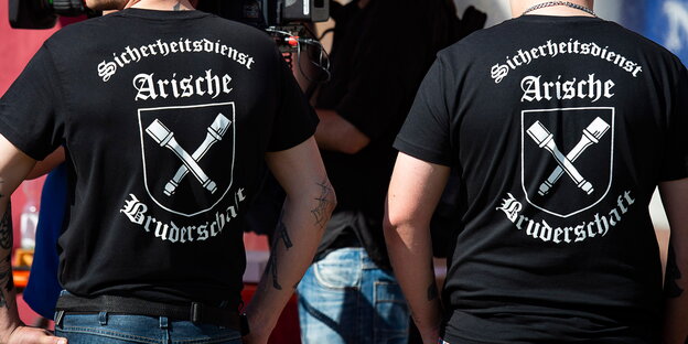die Rücken zweier Männer, auf ihren T-Shirts steht: „Sicherheitsdienst Arische Bruderschaft“ die Rücken zweier Männer, auf ihren T-Shirts steht: „Sicherheitsdienst Arische Bruderschaft“