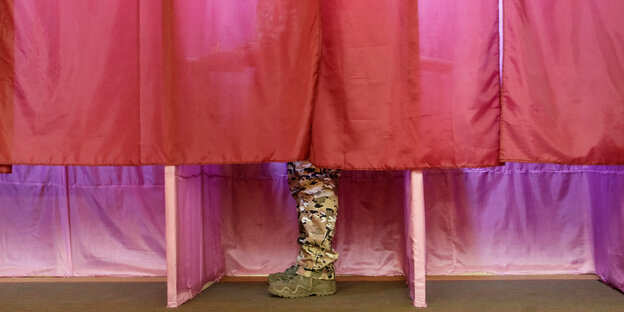 Mit rotem Stoff verhangene Wahlkabinen, in einer sind die Beine eines Mannes in Tarnhose zu sehen Mit rotem Stoff verhangene Wahlkabinen, in einer sind die Beine eines Mannes in Tarnhose zu sehen