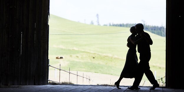 Silhouette eines tanzenden Paares in einer Art Schuppen, im Hintergrund sind grüne Hügel zu sehen Silhouette eines tanzenden Paares in einer Art Schuppen, im Hintergrund sind grüne Hügel zu sehen