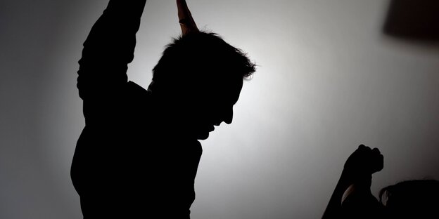 Ein Mann erhebt seine Hände gegen eine andere Person, die ihre Hände sichtbar in die Höhe hält. Im Gegenlicht sind nur die Schatten zu sehen Ein Mann erhebt seine Hände gegen eine andere Person, die ihre Hände sichtbar in die Höhe hält. Im Gegenlicht sind nur die Schatten zu sehen