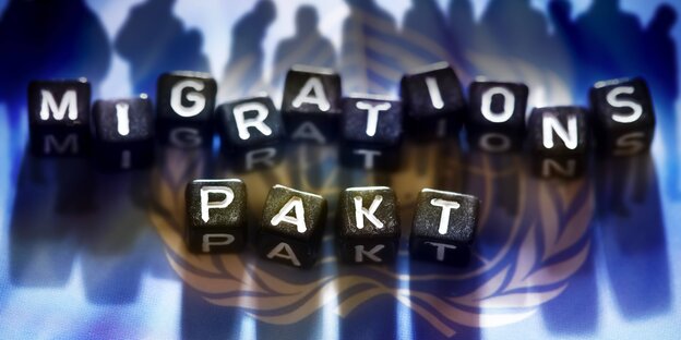 Schwarze Würfel mit weißen Buchstaben ergeben das Wort "Migrationspakt", sie liegen auf der blauen UN-Flagge Schwarze Würfel mit weißen Buchstaben ergeben das Wort "Migrationspakt", sie liegen auf der blauen UN-Flagge
