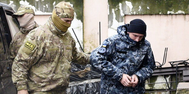 ein vermummter Militär eskortiert einen Mann in blauen Tarnklamotten und in Handschellen ein vermummter Militär eskortiert einen Mann in blauen Tarnklamotten und in Handschellen
