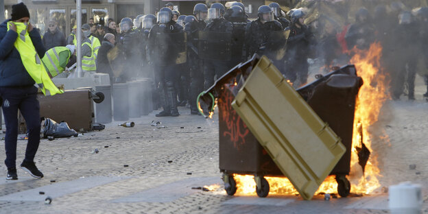 Im Vordergrund eine brennende Mülltonne und ein Protestler, der sich die gelbe Weste vor die Nase hält. Im Hintergrund Polizei Im Vordergrund eine brennende Mülltonne und ein Protestler, der sich die gelbe Weste vor die Nase hält. Im Hintergrund Polizei