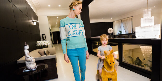 Eine Frau trägt einen Pulli mit der Aufschrift "I'm a Luxury", daneben ein grimmig dreinblickendes Mädchen auf einem Spielzeug-Pony Eine Frau trägt einen Pulli mit der Aufschrift "I'm a Luxury", daneben ein grimmig dreinblickendes Mädchen auf einem Spielzeug-Pony