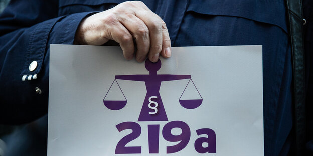 Eine Hand hält ein Schild, auf dem in lila „219a“ steht und eine Waage abgebildet ist Eine Hand hält ein Schild, auf dem in lila „219a“ steht und eine Waage abgebildet ist