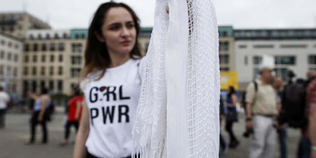 Eine Fraui trägt ein T-Shirt mit dem Aufdruck "GRL PWR" und hält ein weißes Kopftuch in der Hand Eine Fraui trägt ein T-Shirt mit dem Aufdruck "GRL PWR" und hält ein weißes Kopftuch in der Hand