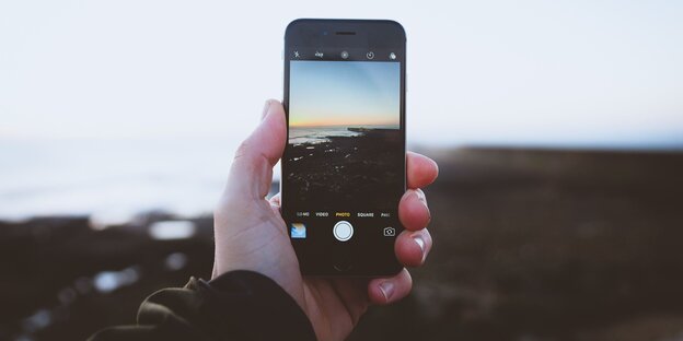 Eine Hand hält ein Smartphone und macht ein Foto vom Meer, das auch im Hintergrund zu sehen it Eine Hand hält ein Smartphone und macht ein Foto vom Meer, das auch im Hintergrund zu sehen it