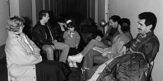 Schwarz-Weiß-Aufnahme: In einem Gang sitzen Personen auf Stühlen, darunter auch Kinder Schwarz-Weiß-Aufnahme: In einem Gang sitzen Personen auf Stühlen, darunter auch Kinder