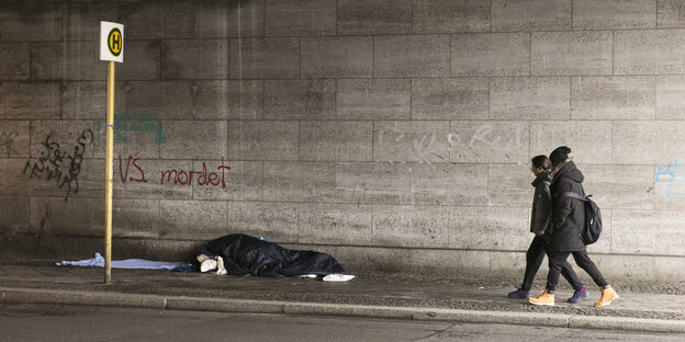Zwei Menschen laufen unter einer Brücke, vor ihnen liegt ein Obdachloser auf dem Boden Zwei Menschen laufen unter einer Brücke, vor ihnen liegt ein Obdachloser auf dem Boden